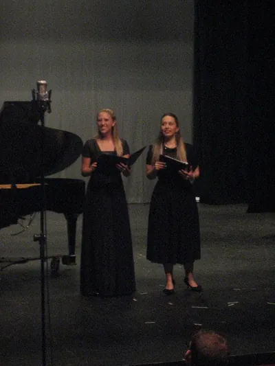 Two singers in long black dresses on a dark stage next to a grand piano